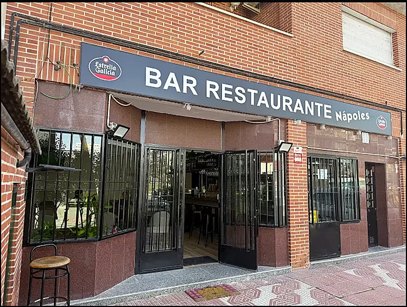 Fachada real de Nápoles Bar y Restaurante en la Calle Nápoles 46: muro de ladrillo, aviso azul marino «BAR RESTAURANTE Nápoles» en letras blancas, el logo de Estrella Galicia, zócalo de granito y un taburete de madera.